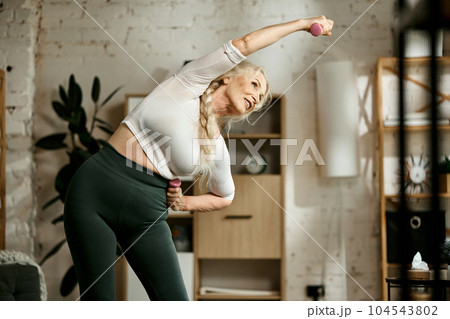 Positive, beautiful, senior woman in comfortable sportswear training, doing warming-up and stretching exercises at home 104543802