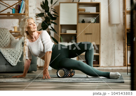 Slim, sportive, beautiful senior woman training at home, keeping body in tone with fitness massage roller. 104543803