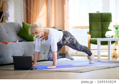 Sportive senior woman looking on laptop on online fitness lessons and training in living room, at home on daytime Sportive senior woman looking on laptop on online fitness lessons and training in living room, at home on daytime 104543829