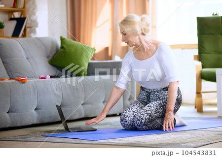 Senior woman training with online fitness lessons at home, doing exercises with trainer via laptop in the morning Senior woman training with online fitness lessons at home, doing exercises with trainer via laptop in the morning 104543831