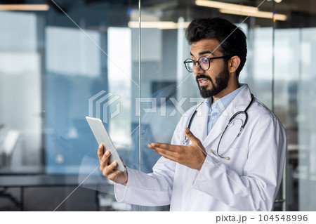 Successful professional Indian doctor working inside modern clinic, man consulting patients remotely, using tablet computer for video call, and long distance communication. Successful professional Indian doctor working inside modern clinic, man consulting patients remotely, using tablet computer for video call, and long distance communication. 104548996