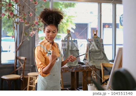 Portrait of creative young woman wearing apron holding palette and brush posing in art studio 104551540
