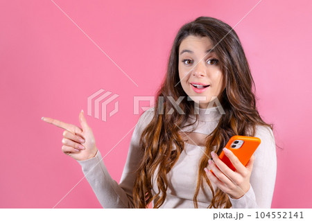 Happy woman with smartphone and shows by finger on the copy space, smiling at the camera and standing against a pink background. Happy woman with smartphone and shows by finger on the copy space, smiling at the camera and standing against a pink background. 104552141