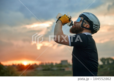 Young sportive man, cyclist in helmet and comfortable clothes standing by the road with bike and drinking water. Sunset 104552862