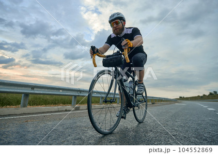 Dynamic image of young bearded man, cyclist in helmet in motion, riding bike on empty road in the evening 104552869