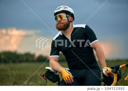 Stylish, bearded, young man in sportswear and helmet, sitting on bike by the road on field in chill summer evening Stylish, bearded, young man in sportswear and helmet, sitting on bike by the road on field in chill summer evening 104552892