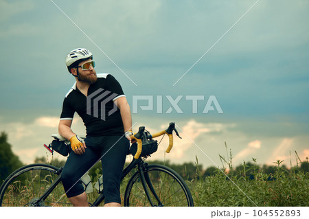 Bearded man in sportswear, glasses and helmet, sitting on bike by the road on field in chill summer evening 104552893