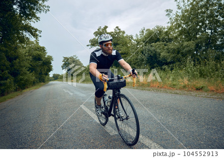 Young bearded man, cyclist in helmet, glasses and uniform riding bike on wet road in cloudy chill evening 104552913