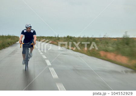 Fast ride. Young bearded man, cyclist in helmet, glasses and uniform riding bike on wet road in cloudy chill evening 104552929