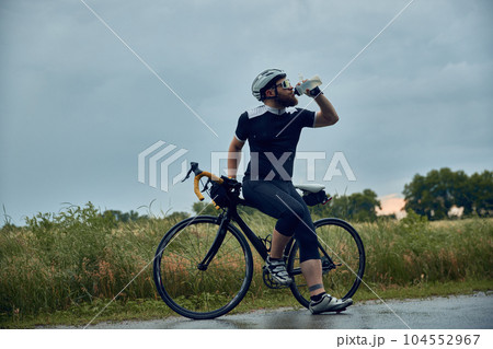 Bearded young man, cyclist in uniform, helmet and glasses standing by the road and drinking water. Cloudy evening Bearded young man, cyclist in uniform, helmet and glasses standing by the road and drinking water. Cloudy evening 104552967