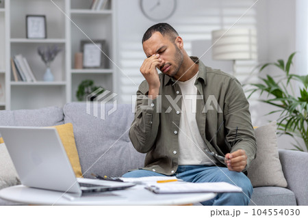 Young African-American man sitting at home tired on couch, resting, rubbing eyes after remote work with laptop and documents. 104554030