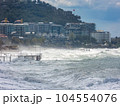 Rough waves crashing into the coast with various buildings situated nearby on the shore 104554076