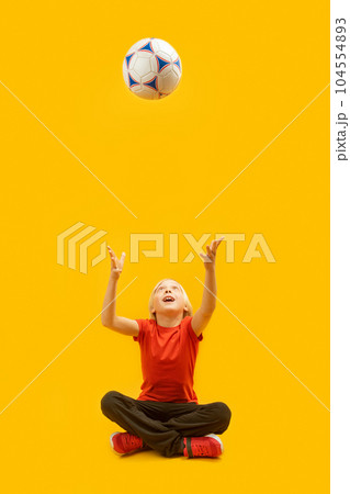 Boy sits cross-legged and tosses soccer ball up in isolation against yellow background. Vertical frame. 104554893