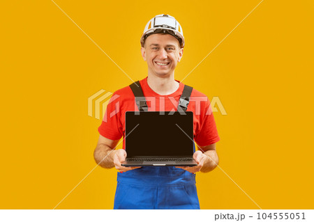 Middle aged man architect engineer wears safety helmet which hand holding laptop with empty screen on yellow studio background. Copy space, mock up. 104555051