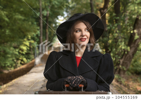 Portrait woman in black hat with brims, classic jaket and gloves rests hands on old suitcase. Forest background Portrait woman in black hat with brims, classic jaket and gloves rests hands on old suitcase. Forest background 104555169