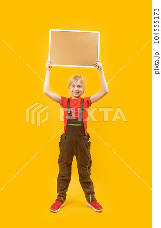 Teenager in overalls holds blank cork board with place for text above his head. Full-length portrait on yellow background. Copy space. 104555173