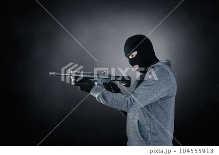Criminal wearing black balaclava and hoodie with assault rifle over dark misty background 104555913