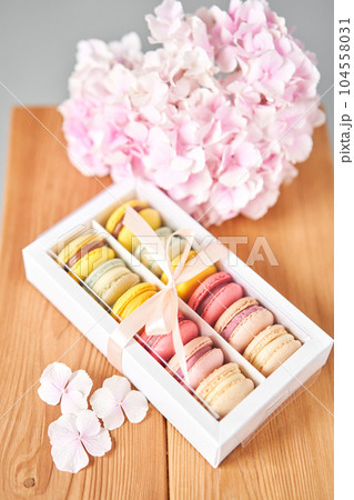 Dessert French Macarons in present box. Sweet dessert on a flowers background. Still life with macaroni cakes and different flower. Delivery concept, coffee and flower shop Dessert French Macarons in present box. Sweet dessert on a flowers background. Still life with macaroni cakes and different flower. Delivery concept, coffee and flower shop 104558031