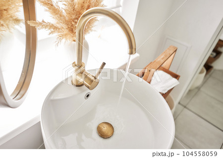 A beautiful sink with a golden faucet next to an oval mirror and a shelf with hand towels. Close-up of an elegant golden faucet in the bathroom sink next to stylish decorations. A beautiful sink with a golden faucet next to an oval mirror and a shelf with hand towels. Close-up of an elegant golden faucet in the bathroom sink next to stylish decorations. 104558059