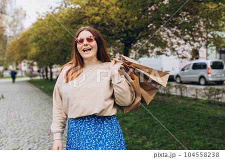 Happy young redhead woman holding shopping paper bags in hands, walking at city street, black friday. Urban lifestyle concept. Shopping banner, for mockup 104558238
