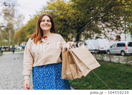 Happy young redhead woman holding shopping paper bags in hands, walking at city street, black friday. Urban lifestyle concept. Shopping banner, for mockup 104558239