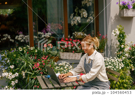 Beautiful flower shop owner wearing apron working on laptop in her store Beautiful flower shop owner wearing apron working on laptop in her store 104560453