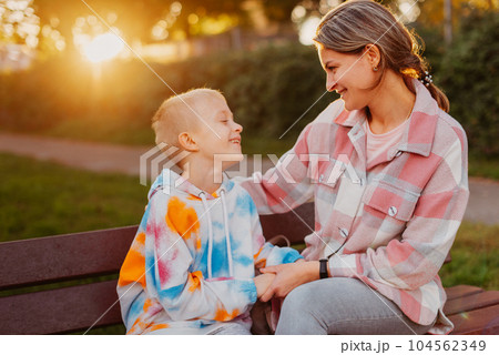 mother and son sit on a park bench in the rays of the setting sun. the concept of a family. Mother's Day. beautiful girl (mother) with a boy (son) in the park in the park are sitting on a bench at mother and son sit on a park bench in the rays of the setting sun. the concept of a family. Mother's Day. beautiful girl (mother) with a boy (son) in the park in the park are sitting on a bench at 104562349