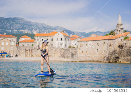 Young women Having Fun Stand Up Paddling in blue water sea in Montenegro. Against the backdrop of the Old Town of Budva. SUP 104568552