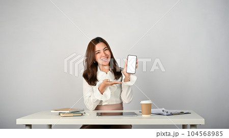 A beautiful Asian businesswoman sits at her desk and shows her phone white screen mockup 104568985