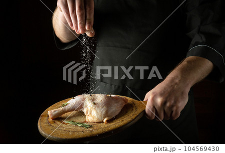 Chicken leg on a cutting board in the chef's hand. The cook salts the chicken leg before cooking or roasting Chicken leg on a cutting board in the chef's hand. The cook salts the chicken leg before cooking or roasting 104569430