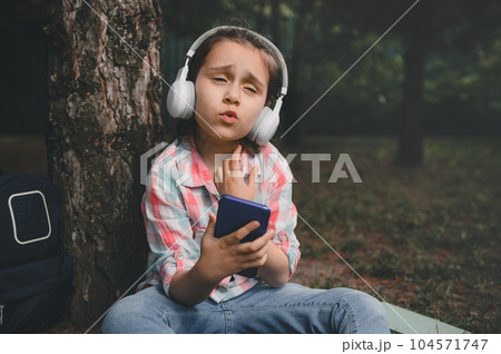 Caucasian little child girl listening to music on wireless headphones, singing song, holding smart mobile phone outdoors Caucasian little child girl listening to music on wireless headphones, singing song, holding smart mobile phone outdoors 104571747
