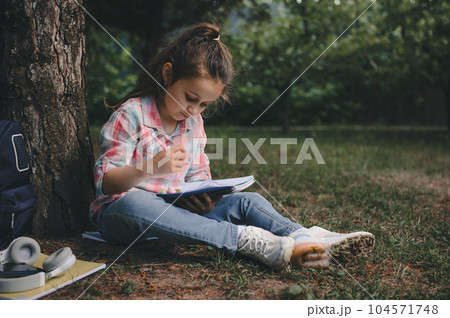 Cute little child girl, primary school student writing on copybook, doing homework outdoors. School and children concept Cute little child girl, primary school student writing on copybook, doing homework outdoors. School and children concept 104571748