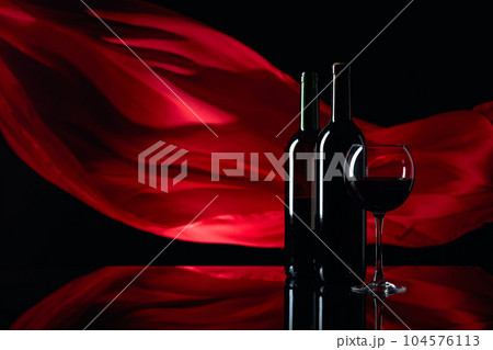 Wineglass and bottles of red wine on a black reflective background. Red satin curtain flutters in the wind. Wineglass and bottles of red wine on a black reflective background. Red satin curtain flutters in the wind. 104576113