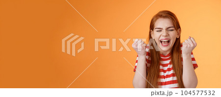 I did it. Charming lucky determined redhead female achieve success, fist pump celebrating, close eyes yell yes yeah, hooray gesture, triumphing victory, very good news, stand orange background 104577532