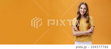 Portrait of kind and friendly charming young female student in casual t-shirt with wavy natural long hair holding hands crossed on chest smiling broadly and carefree at camera over orange background Portrait of kind and friendly charming young female student in casual t-shirt with wavy natural long hair holding hands crossed on chest smiling broadly and carefree at camera over orange background 104577788