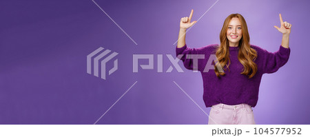 Scroll up and see cool copy space. Portrait of happy attractive and friendly young ginger woman with freckles in warm purple sweater raising hands pointing upwards as showing place Scroll up and see cool copy space. Portrait of happy attractive and friendly young ginger woman with freckles in warm purple sweater raising hands pointing upwards as showing place 104577952