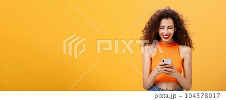 Stylish young attractive woman with curly hairstyle, nose ring and red lipstick picking song, typing in smartphone listening music in wireless earphones smiling at device screen over orange wall 104578017
