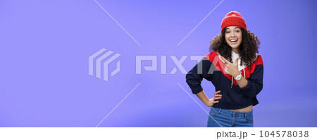 Stylish and sociable friendly-looking attractive woman with curly hair in warm beanie pointing at upper left corner laughing from joy and amazement having fun posing happily over blue background 104578038
