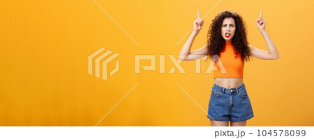 Indoor shot of displeased female client with curly stylish hairstyle red lipstick in cool cropped top pointing up demanding explanation with confused and dissatisfied expression over orange background 104578099