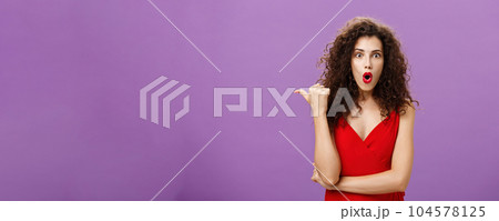 Curious and questioned amazed woman with curly hairstyle in stylish red dress folding lips in wow sound pointing left with thumb asking question about stunning and thrilling scene over purple wall Curious and questioned amazed woman with curly hairstyle in stylish red dress folding lips in wow sound pointing left with thumb asking question about stunning and thrilling scene over purple wall 104578125