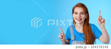 Close-up cheerful carefree redhead dancing girl pointing up, raising fingers, smiling amused, having fun, stand blue background entertained, enjoy music party, disco mood, carefree emotions concept 104578148