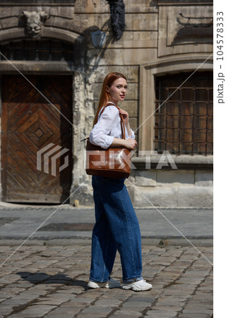 photo of a woman wearing blue jeans and white blouse with a brown leather handbag . 104578333
