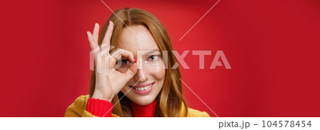 Zero worries only happiness. Portrait of charming cute and carefree redhead female with freckles and blue eyes smiling delighted showing okay gesture on eye and peeking at camera over red background 104578454