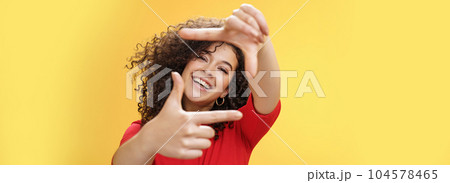 Portrait of optimistic happy and creative female student imaging her new apartment as extending hands and showing frames gesture smiling through it at camera amused and carefree over yellow wall 104578465