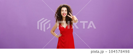 Studio shot of friendly gorgeous and stylish young european woman with cute curly hairstyle red lipstick and dress showing peace gesture at camera and smiling broadly over purple background 104578498