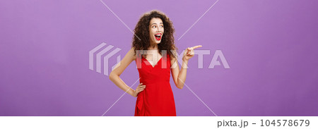 Amused attractive european female with evening makeup in luxurious red dress and curly hairstyle laughing out loud entertained and happy pointing and gazing left intrigued over purple background 104578679