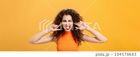 Waist-up shot of irritated bothered stylish woman with curly hair and red lipstick squinting clenching teeth waiting for loud bang covering ears with index fingers being annoyed of noisy neighbours Waist-up shot of irritated bothered stylish woman with curly hair and red lipstick squinting clenching teeth waiting for loud bang covering ears with index fingers being annoyed of noisy neighbours 104578683