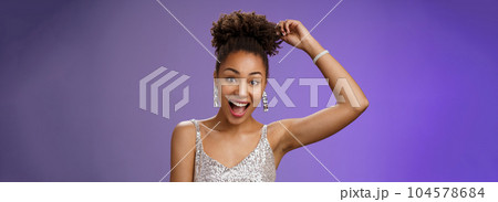 Close-up excited good-looking african-american curly-haired woman in trendy silver glittering dress touching afro curly smiling gladly having fun dancing amused standing entertained blue background 104578684