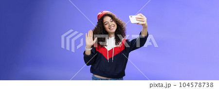 Studio shto of stylish cute female vlogger with curly hair in winter red hat smiling and waving hello at smartphone camera as recording video, making interesting content to post online over blue wall 104578738