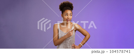 Curious unsure african-american attractive elegant young woman in silver glittering evening dress look pointing left intrigued discuss strange person gaze doubtful uncertain, blue background Curious unsure african-american attractive elegant young woman in silver glittering evening dress look pointing left intrigued discuss strange person gaze doubtful uncertain, blue background 104578741
